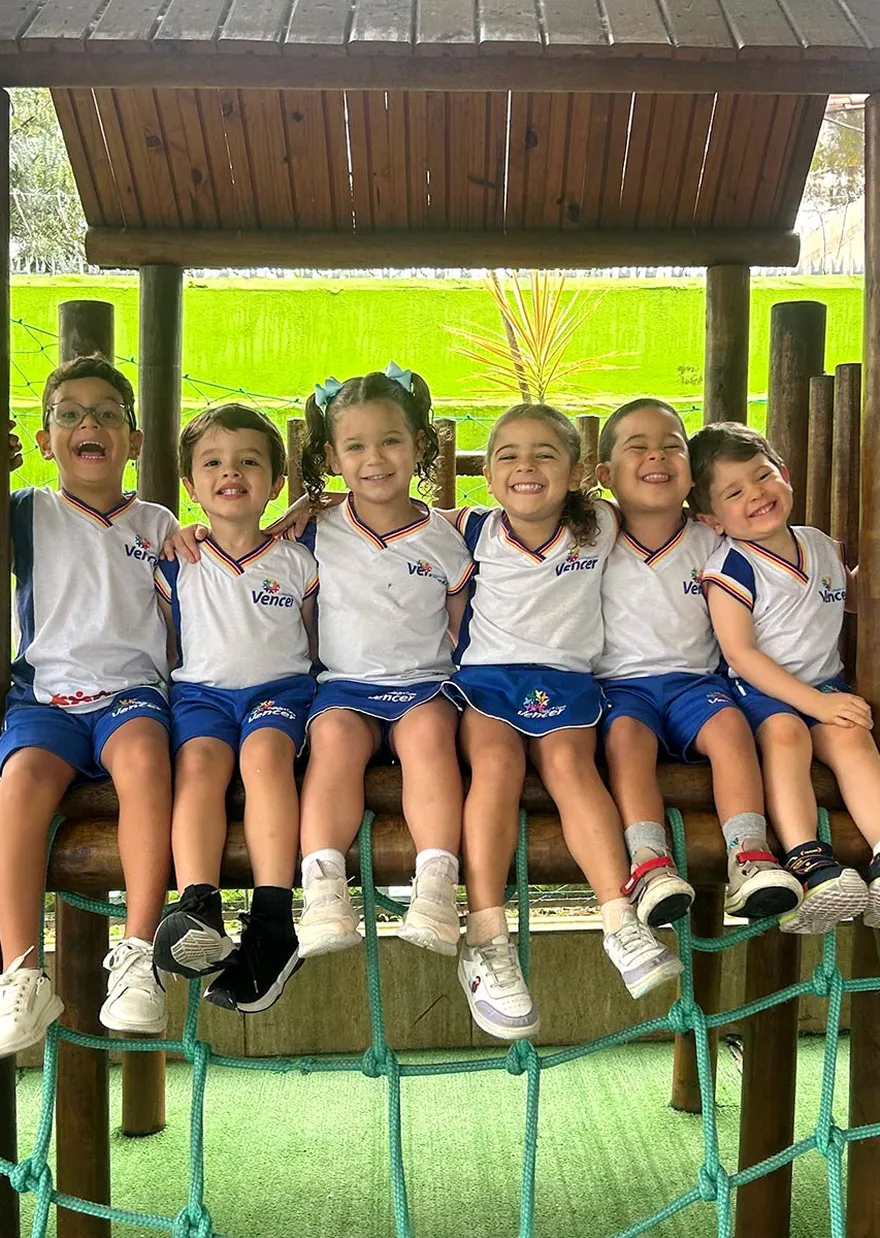 Seis alunos do Colégio Vencer sorrindo, sentados juntos no parquinho da escola